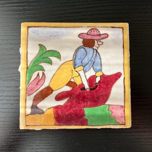 May include: A square ceramic tile with a hand-painted design. The image depicts a person in a blue shirt and pink hat riding a red creature. The tile has a yellow border and green and pink floral accents.