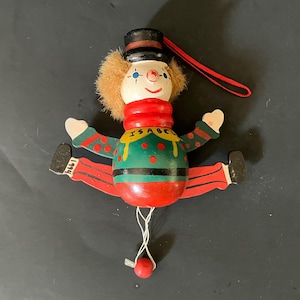 May include: A vintage wooden clown ornament with a red and green color scheme. The clown has a black top hat, a fluffy brown wig, and a painted face. The word "ISABEL" is painted on the clown's chest. The clown's legs are spread, and it has a red string for hanging.
