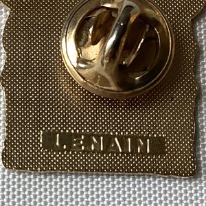 Op de afbeelding: Gouden metalen pin met een getextureerd oppervlak en een pin achterkant. De pin heeft een ontwerp van een gebouw met de tekst "LE NAIN" op de achterkant.