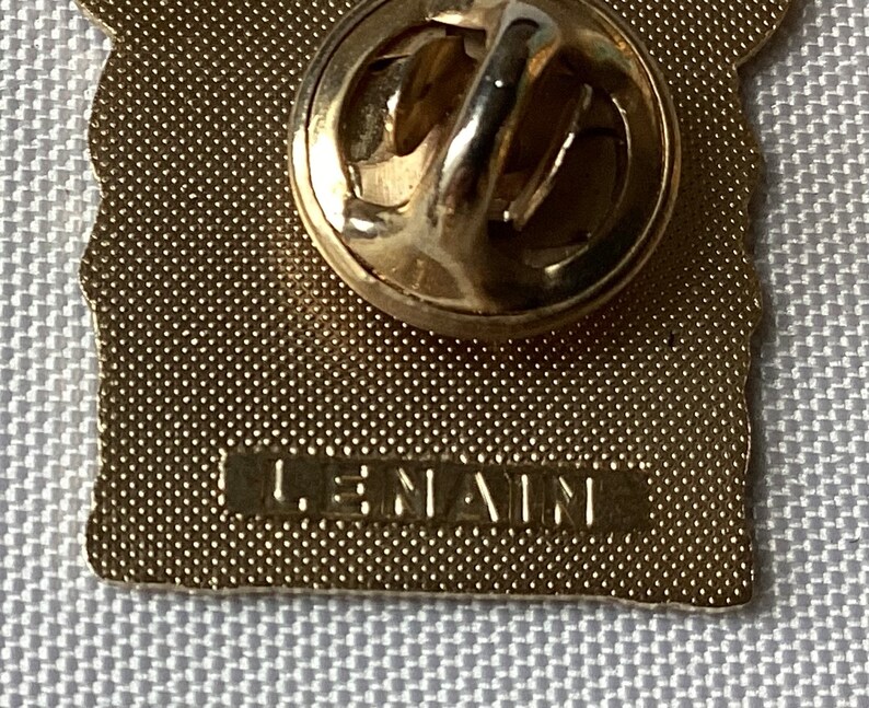 Op de afbeelding: Gouden metalen pin met een getextureerd oppervlak en een pin achterkant. De pin heeft een ontwerp van een gebouw met de tekst "LE NAIN" op de achterkant.