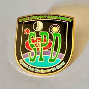 May include: A shield-shaped metal pin with a black background featuring the green text SPD, a red oval, a purple arc, and a blue horizon. The top text reads SPACE PRODUCT DEVELOPMENT and the bottom text reads Space For Business to Grow.