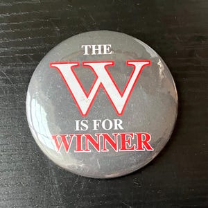May include: A round, gray button with the text "THE W IS FOR WINNER" in white and red lettering. The large letter "W" is in the center, with a red outline. The button has a glossy finish.