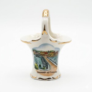 May include: A white ceramic basket with a gold-trimmed handle and rim. The front features a colorful illustration of a waterfront cityscape with a bridge and the text Souvenir of Curacao printed in black script at the base of the illustration.