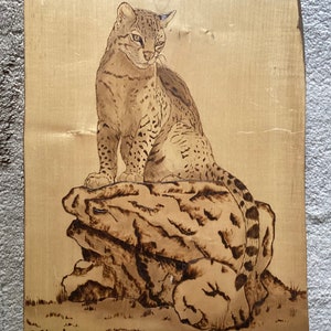 May include: A woodburned image of a spotted cat sitting on a rock. The image is signed "D. Strack '88" in the bottom left corner.