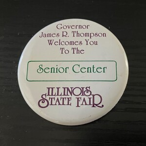 May include: A white button with text that reads "Governor James R. Thompson Welcomes You To The Senior Center" and "Illinois State Fair" in purple and green lettering.