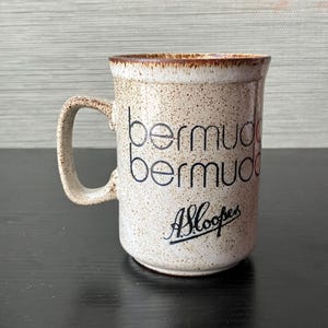 May include: A speckled ceramic mug with the word "Bermuda" in black script repeated twice, and the signature "A.Sloopes" in cursive. The mug has a brown rim and handle, and is set against a neutral background.
