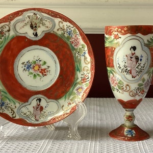 May include: A vintage Japanese teacup and saucer set with a red and white floral design. The cup has a handle and a footed base. The saucer has a raised rim and a floral design.
