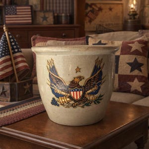 May include: A beige stoneware crock with a patriotic eagle emblem. The eagle has blue and gold wings, a red, white, and blue shield, and is surrounded by olive branches. A star is above the eagle. The crock is on a wooden table.