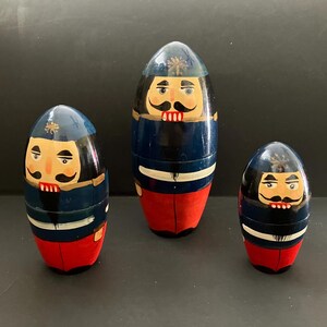 May include: Three wooden nesting dolls, each painted to resemble a soldier. The dolls are blue, red, and white, with detailed facial features including mustaches. The largest doll is approximately 6 inches tall.