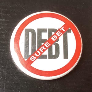 May include: A round button with a white background and a red border. The word "DEBT" is printed in large, bold, black letters. A red diagonal line crosses out the word "DEBT", with the words "SURE BET" printed diagonally across the line.