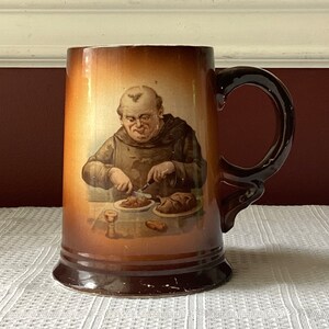May include: A brown ceramic beer stein with a handle. The stein features a painted image of a man in a brown robe holding a knife and cutting a piece of meat. The man is sitting at a table with a plate of food in front of him.