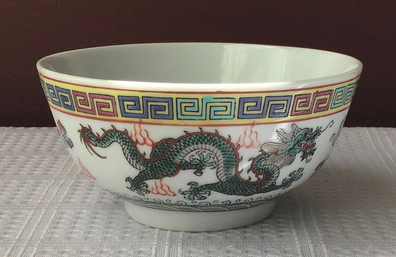 May include: White porcelain bowl with a green and blue dragon design. The bowl has a yellow, blue, and red border with a repeating geometric pattern.