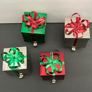May include: Four small, square gift boxes with metallic finishes. Two are silver, one is green, and one is red. Each box is adorned with a large, decorative bow in a contrasting color. The boxes have a small metal clasp at the bottom.