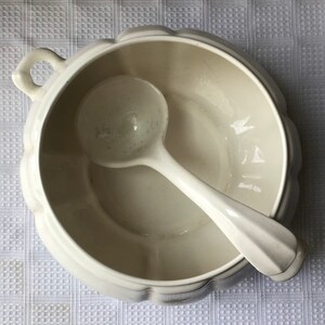 May include: White ceramic soup tureen with a matching ladle. The tureen has a fluted rim and a handle on the side. The ladle is resting inside the tureen.