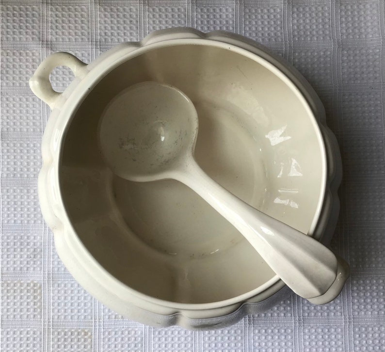 May include: White ceramic soup tureen with a matching ladle. The tureen has a fluted rim and a handle on the side. The ladle is resting inside the tureen.