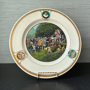 May include: A round ceramic decorative plate featuring a central illustration of a historical battle scene with figures in period clothing. The rim is cream-colored with gold trim, a portrait medallion at the top, and two heraldic crests on the sides.