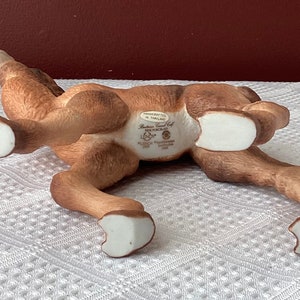 May include: A brown ceramic figurine of a reclining deer with white hooves. The underside of the deer has text that reads "Handcrafted in Thailand" and "Fine Porcelain". The deer is lying on a white patterned surface.