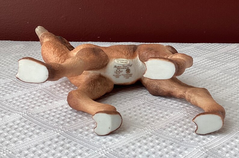 May include: A brown ceramic figurine of a reclining deer with white hooves. The underside of the deer has text that reads "Handcrafted in Thailand" and "Fine Porcelain". The deer is lying on a white patterned surface.
