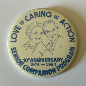 May include: A round off-white pinback button featuring a blue line-art illustration of two people. The text reads LOVE IS CARING IN ACTION, 10th ANNIVERSARY, 1974-1984, and SENIOR COMPANION PROGRAM in blue capital letters around the central image.