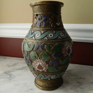 K&ouml;nnte beinhalten: Eine Messingvase mit Cloisonn&eacute;-Design. Die Vase ist mit floralen Mustern in Blaut&ouml;nen, Gr&uuml;n, Rosa und Rot verziert. Die Vase hat einen runden K&ouml;rper und eine ausgestellte Lippe.