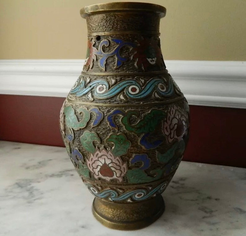 K&ouml;nnte beinhalten: Eine Messingvase mit Cloisonn&eacute;-Design. Die Vase ist mit floralen Mustern in Blaut&ouml;nen, Gr&uuml;n, Rosa und Rot verziert. Die Vase hat einen runden K&ouml;rper und eine ausgestellte Lippe.