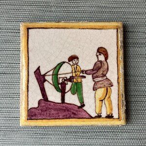 May include: A hand-painted ceramic tile depicting two figures, one operating a wheel and the other standing beside it. The tile has a yellow border and a white background with colorful details.