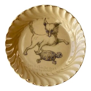 May include: A cream-colored ceramic plate with a gold rim and a ruffled edge. The plate features a black and white illustration of a dog standing on a turtle. The text "Den BT Tox, he grab him by detail" is below the illustration.