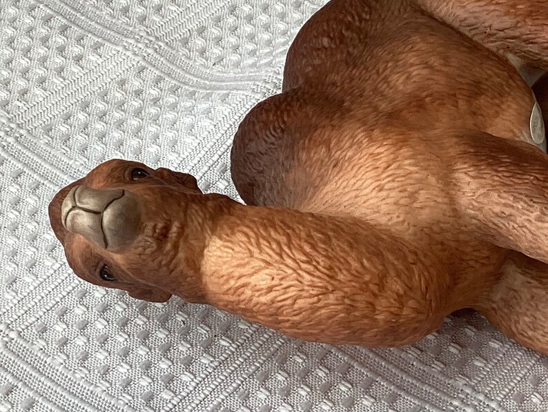 May include: A ceramic camel figurine in shades of brown, with detailed texture and a serene expression. The camel is lying down, with its head turned towards the viewer. The figurine is likely a decorative item.