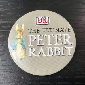 May include: A round button featuring Peter Rabbit in a blue jacket. The button has the text "THE ULTIMATE PETER RABBIT" and the DK logo. The background is a light green color.