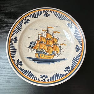 May include: A decorative ceramic plate featuring a hand-painted sailing ship with orange sails and a blue hull. The plate has a cream-colored background with a blue and yellow border. The plate is approximately 10 inches in diameter.