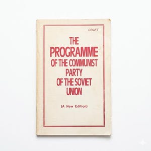 May include: A rectangular book with a cream-colored cover and a thin red border. The text in red reads: DRAFT, THE PROGRAMME OF THE COMMUNIST PARTY OF THE SOVIET UNION, and (A New Edition). The cover has a matte texture and shows signs of age.