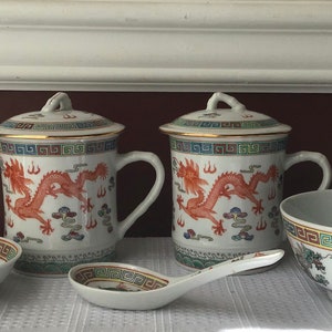 May include: Two white ceramic mugs with gold trim and red dragon designs. The mugs have lids and are sitting on a white surface. There is a small white saucer and a white bowl with a dragon design on it.