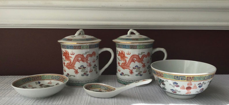 May include: Two white ceramic mugs with gold trim and red dragon designs. The mugs have lids and are sitting on a white surface. There is a small white saucer and a white bowl with a dragon design on it.