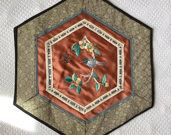 May include: A hexagonal table runner with a brown background and a gold and blue border. The center features an embroidered design of a bird perched on a branch with flowers and a butterfly.