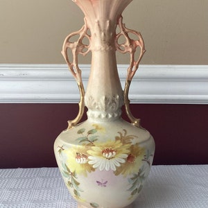 May include: A vintage porcelain vase with ornate handles and a floral design. The vase is decorated with yellow and white flowers and has a pink and gold glaze.
