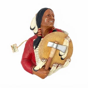 May include: A wall-mounted Bossons chalkware bust depicting a figure with long black hair, wearing a red shirt, holding a tan circular shield with white feathers and a silver-headed tomahawk. A small paper tag labeled Bossons is attached by a string.