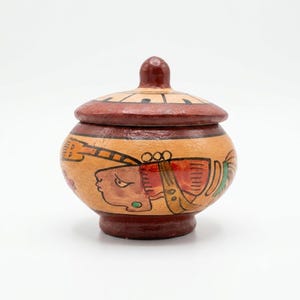 May include: A round ceramic lidded jar featuring a tan base color with hand-painted illustrations of a profile figure in red, black, and green. The lid and base are finished in a deep reddish-brown tone, with a small knob handle on the lid.