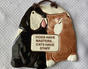 May include: A ceramic wall hanging featuring a black dog and a brown cat. The dog and cat are facing each other and have a sign that reads "Dogs have masters... Cats have staff".