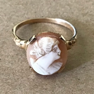 May include: A gold ring with a cameo of a woman's profile. The cameo is made of white shell and is set in a gold bezel. The ring is a delicate design with a simple band.