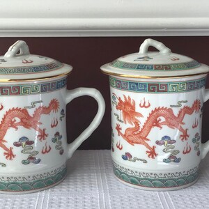 May include: Two white ceramic teacups with lids, featuring a red dragon design and gold trim. The cups have a traditional Chinese pattern and are perfect for serving tea.