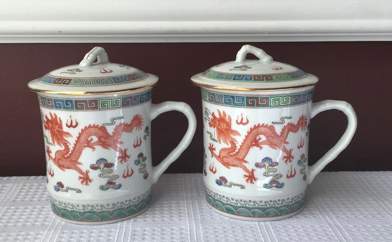 May include: Two white ceramic teacups with lids, featuring a red dragon design and gold trim. The cups have a traditional Chinese pattern and are perfect for serving tea.
