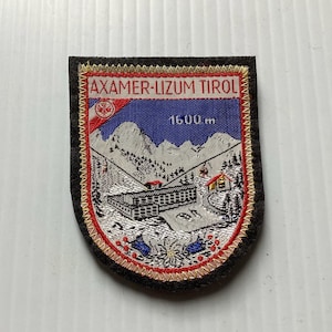 May include: Embroidered patch with the text "AXAMER-LIZUM TIROL" and "1600 m." The patch features a mountain scene with a building, trees, and a ski lift against a blue sky. The patch has a black border with red and gold accents.