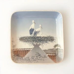 May include: A square porcelain plate with a blue and white design of storks in a nest. The storks are white with black wings and beaks. The nest is made of twigs and is surrounded by a light blue background.