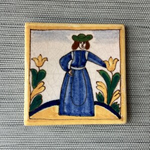 May include: A hand-painted ceramic tile featuring a woman in a blue dress and a green hat, standing in a garden with yellow flowers. The tile has a yellow border and a white background.