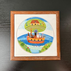 May include: A framed tile featuring a colorful illustration of Noah's Ark. The ark is orange and filled with animals, sailing on blue water. A green tree and house are above the ark, with a brown wooden frame.