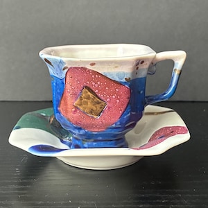 May include: A ceramic teacup and saucer set. The cup and saucer are white with blue, red, and green abstract designs. The cup has a blue handle and a red and brown square design.