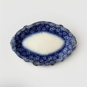 May include: Oval-shaped antique serving platter with a white center and a blue floral pattern border. The rim features a detailed, ornate design. The platter is displayed on a black metal stand, showcasing its vintage aesthetic.