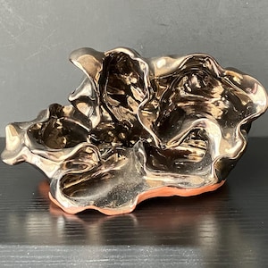 May include: A metallic gold-colored decorative bowl with a unique, organic shape. The bowl has a rippled, textured surface and a reddish-brown base. The reflective surface creates highlights and shadows, enhancing its sculptural form. The bowl sits on a dark surface.