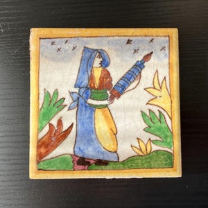 May include: A hand-painted ceramic tile depicting a woman in a blue dress holding a rolling pin. The tile has a yellow border and a white background with green and yellow floral designs.