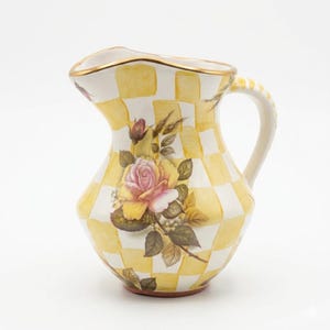 May include: A ceramic pitcher featuring a yellow and white checkered pattern with a central floral design of a pink and yellow rose with green leaves. The rim of the pitcher is accented with a thin gold-colored trim, and it has a curved handle.